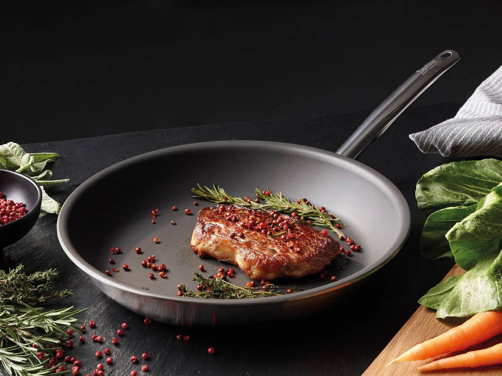 Spring Brigade Ultimate Koekenpan - 20 cm, op een donkere ondergrond met een gebakken steak, roze peper, rozemarijn en verse groenten eromheen