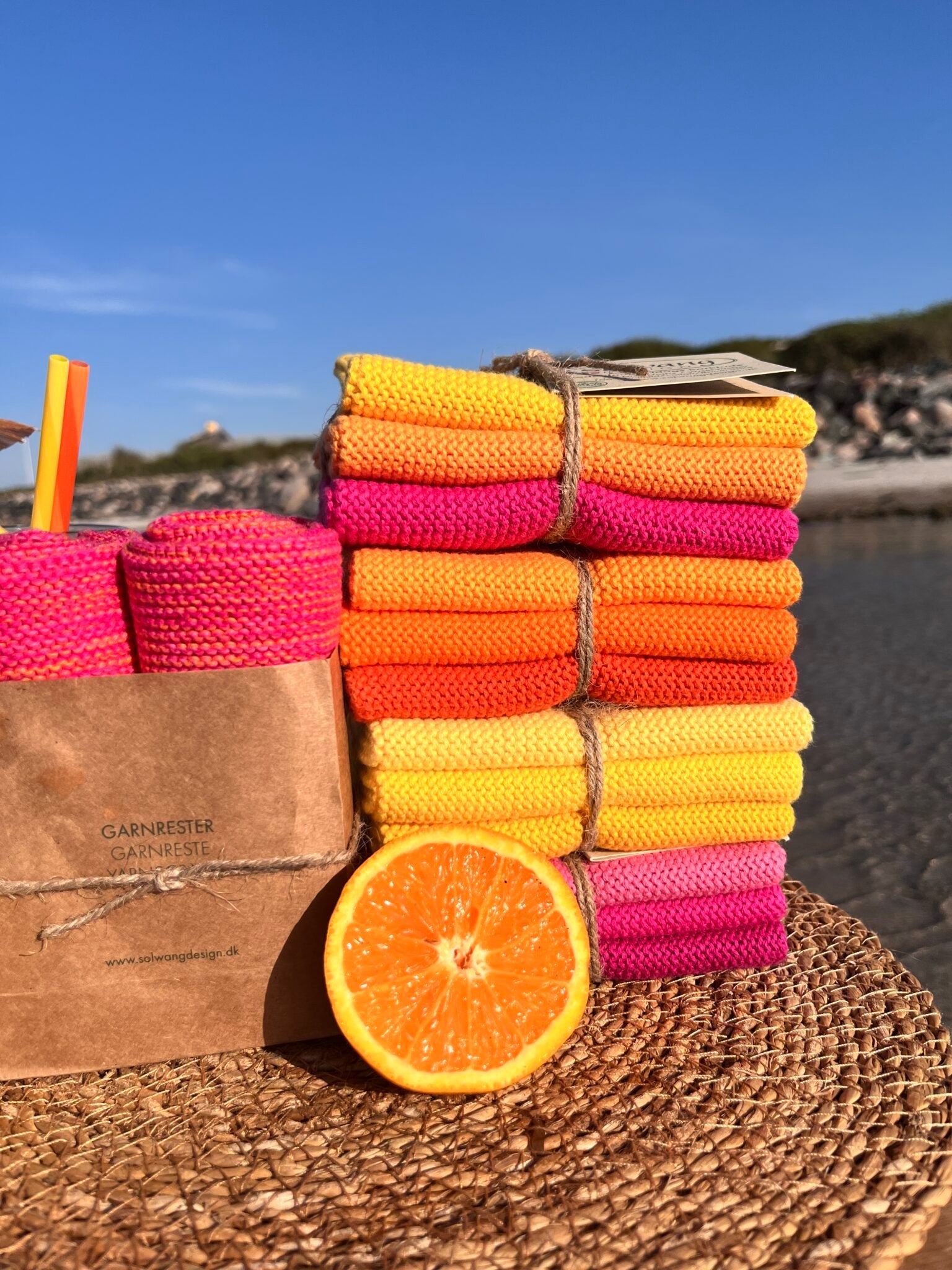Solwang Design Vaatdoekjes set van 3 - Oranje Combi sfeerfoto buiten aan het water met meerdere gebonden setjes Solwang-doekjes in oranje, geel en roze/framboos tinten op elkaar gestapeld, een doorgesneden sinaasappel op de voorgrond en een kraftpapieren zakje met opschrift Garnrester ernaast.