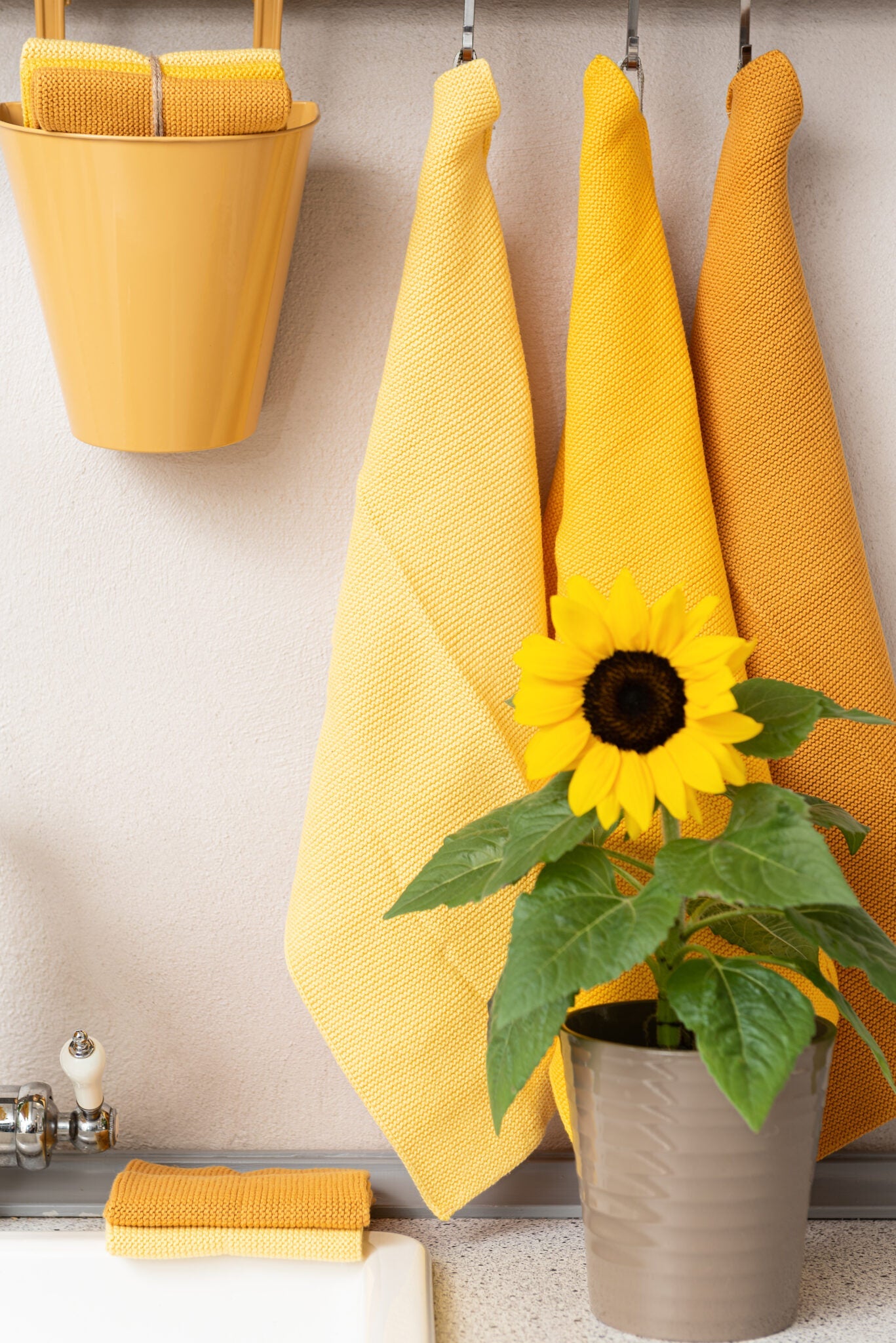 Solwang Design Vaatdoekjes set van 3 - Donkergeel Combi sfeerfoto met drie gele/okergele doekjes hangend aan haken tegen een muur, met een zonnebloem in een plantenbak ervoor, een geel emmertje met opgevouwen doekjes ernaast en nog een opgevouwen doekje op de vensterbank onderin beeld.