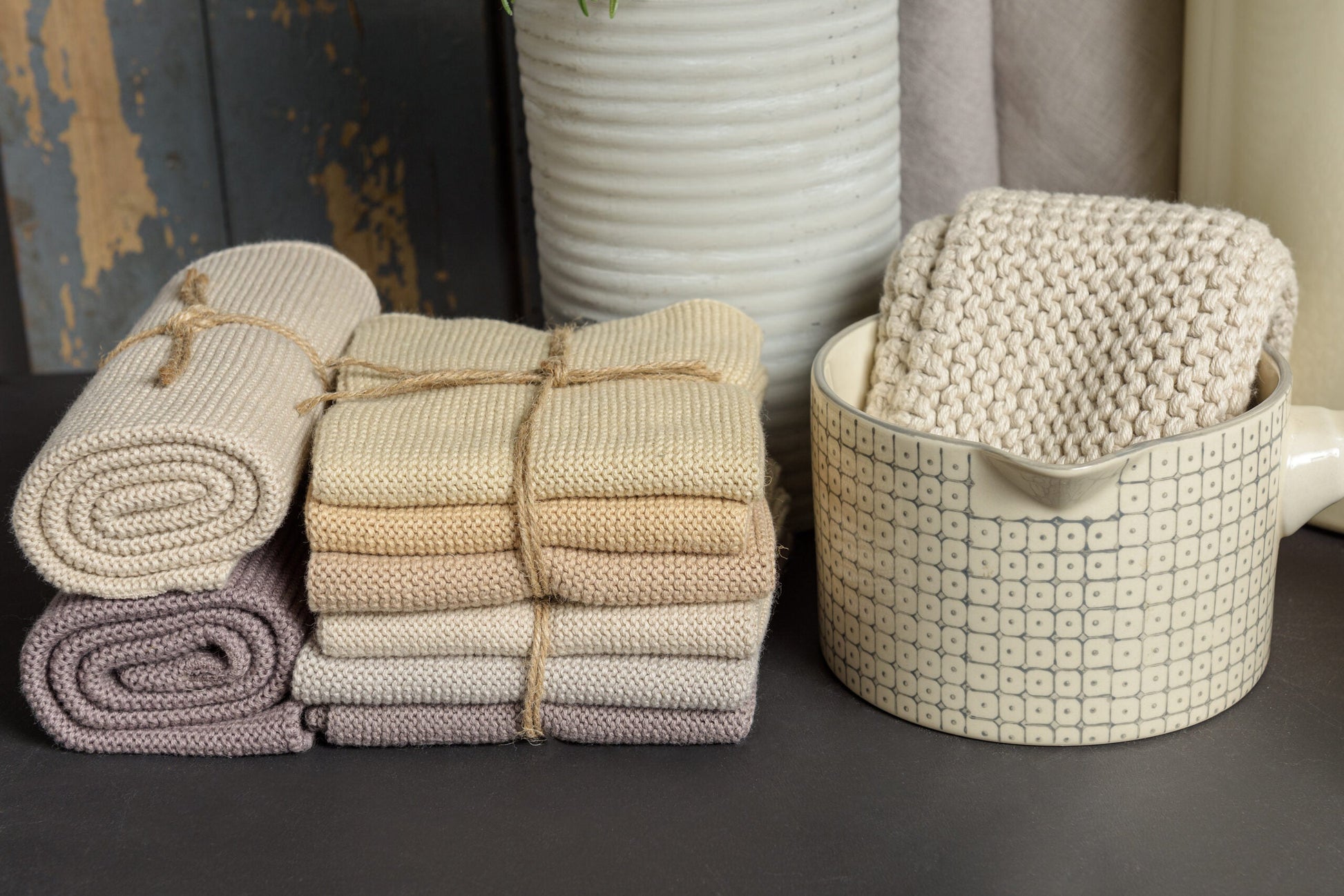 Solwang Design Vaatdoekjes set van 3 - Zand Combi sfeerfoto met een opgerold doekje in zandtint, een gestapeld setje opgevouwen doekjes in zand-, beige- en lichtpaarse tinten en een gevouwen doekje in een keramische kom, tegen een rustieke houten achtergrond met een witte geribbelde vaas.