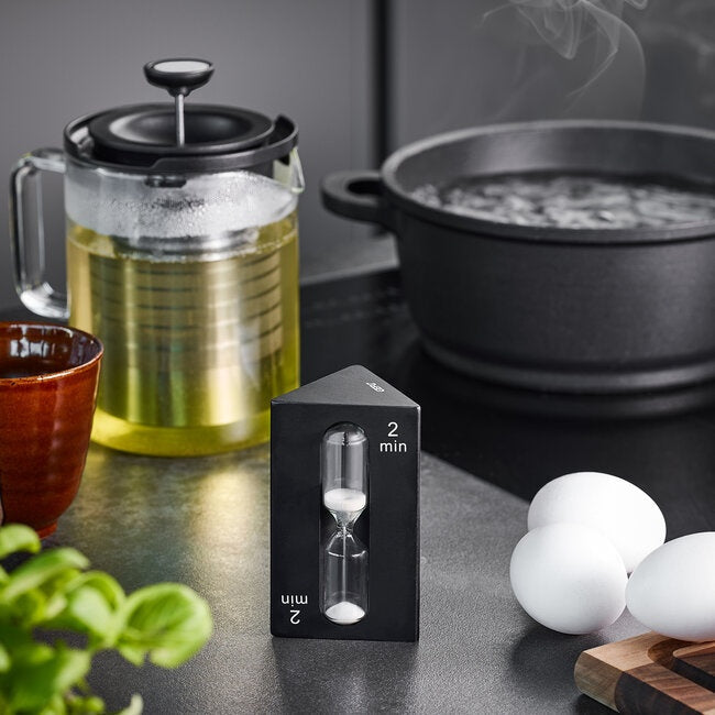 GEFU Zandloper Sando, de kleinere zandloper van 2 minuten op het aanrecht naast een cafetière en een pan met kokend water, met eieren op de achtergrond