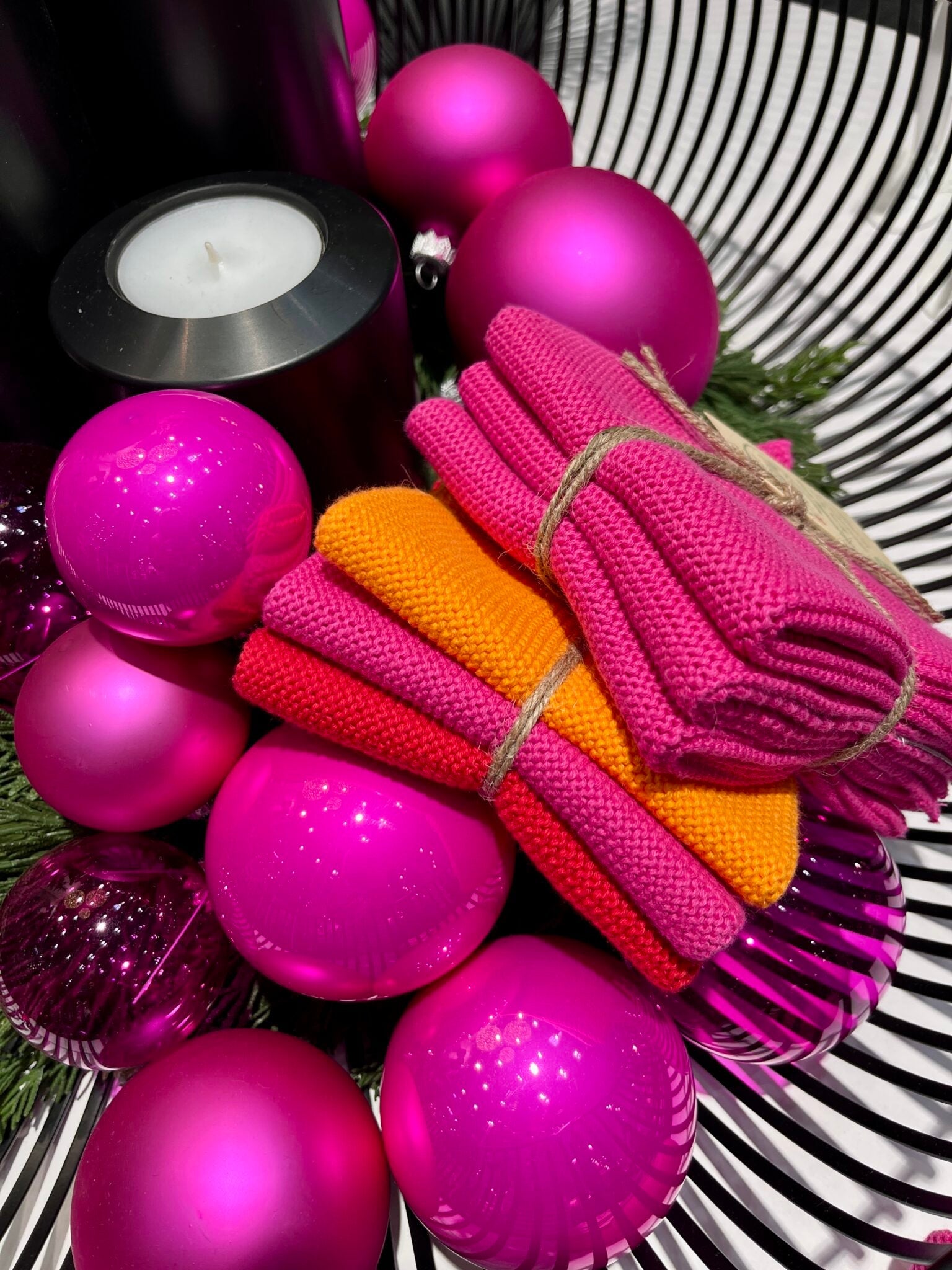 Solwang Design Vaatdoekjes set van 3 - Oranje/Roze/Rood kerstachtige sfeerfoto met fuchsia glazen kerstballen, een zwarte kandelaar met brandend waxinelichtje en dennengroen, waartussen een gebonden setje roze/fuchsia vaatdoekjes en een los gestapeld setje in oranje, roze en rood liggen.