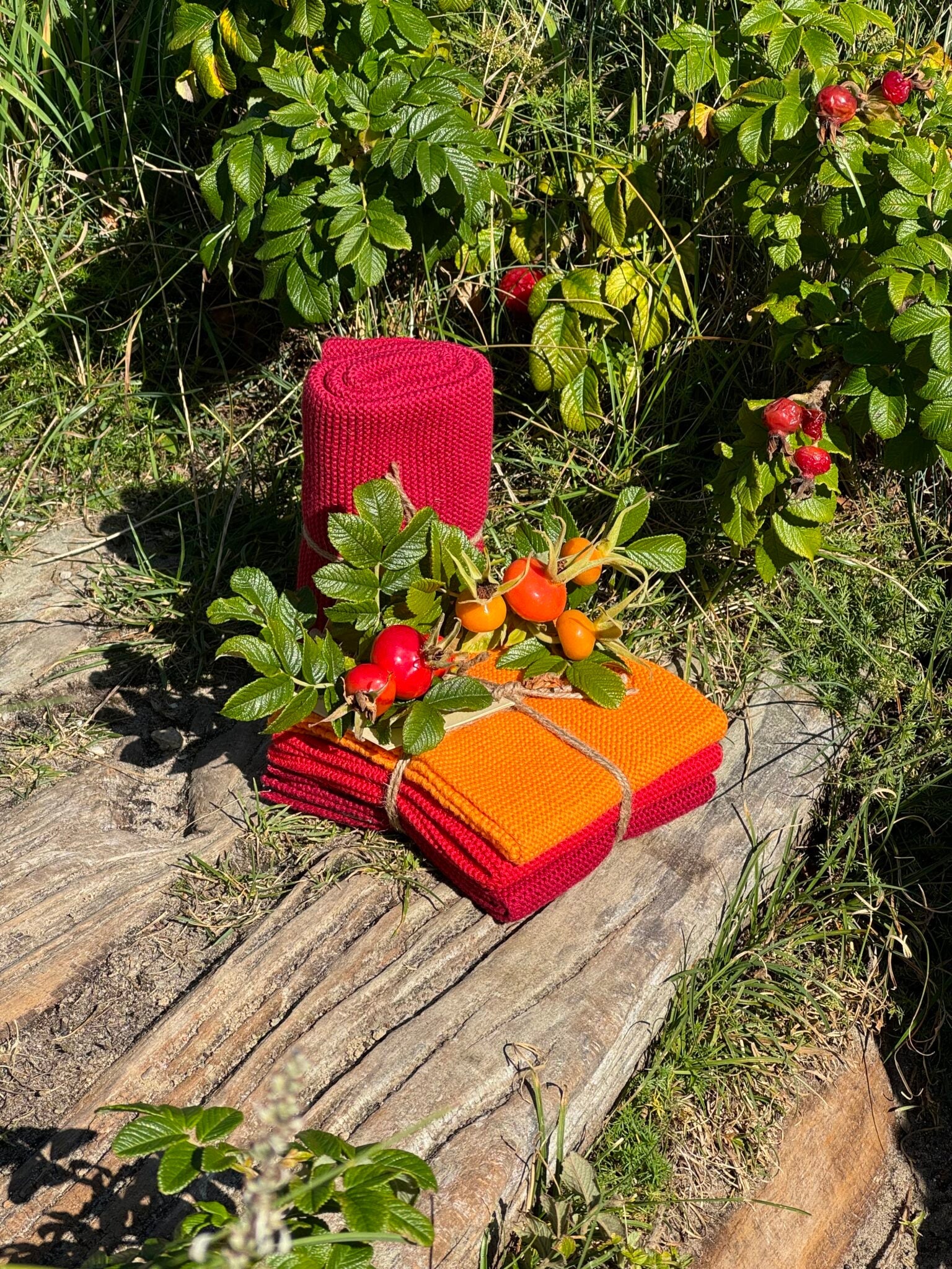 Solwang Design Vaatdoekjes set van 3 - Oranje/Rood Combi lifestylefoto tussen een rozenbottelstruik: een rood doekje staat rechtop opgerold, terwijl een rood en een oranje doekje gestapeld en met touw samengebonden liggen op een houten plank, versierd met een takje bladeren en rozenbottels.