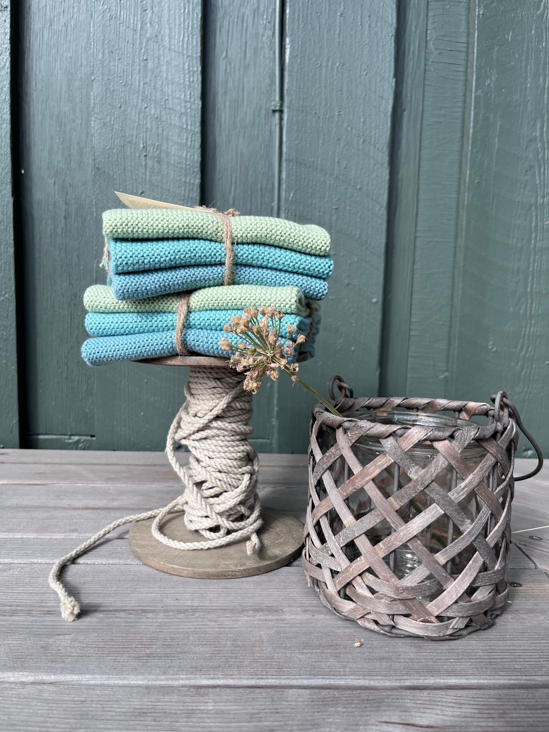 Solwang Design Vaatdoekjes set van 3 - Zeeschuim Combi lifestylefoto met de doekjes in mint, turquoise en blauw gestapeld in twee setjes op een met touw omwikkelde houten klos, met een gedroogde sierui-bloem ertussen gestoken en een gevlochten rieten lantaarn ernaast tegen een grijsgroen geschilderde houten achtergrond.