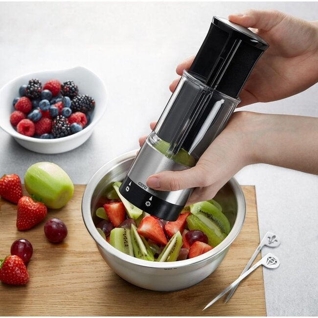 GEFU Groente- en Fruitsplitter Flexicut, twee handen die het toestel gebruiken om kiwi in stukjes te snijden boven een kom met aardbeien, bosbessen, druiven en appel op een houten snijplank