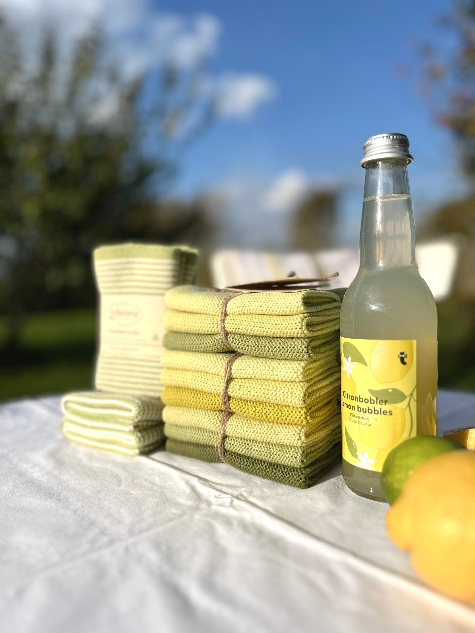 Solwang Design Vaatdoekjes set van 3 - Pistache Combi lifestylefoto op een wit tafelkleed buiten: een stapel geel/groene doekjes samengebonden met touw staat naast een flesje limonade met het etiket Citronbobler Lemon Bubbles en citrusvruchten, met een tuin op de achtergrond.