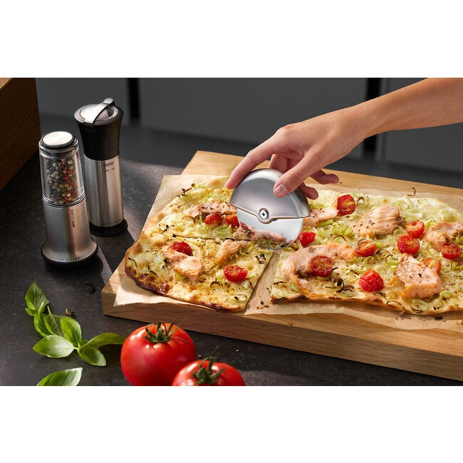 GEFU Pizzasnijder Monza, een hand snijdt met de ronde RVS pizzasnijder door een pizza met zalm en cherrytomaatjes op een houten plank