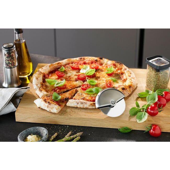 GEFU Pizzasnijder Monza op een houten plank naast een pizza met tomaat en basilicum, omringd door olijfolie, asperges en cherrytomaatjes