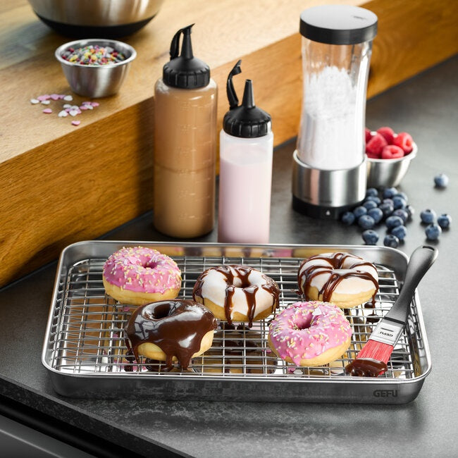 GEFU Bakplaat Variado - 31,5 cm met rooster gevuld met versierde donuts met chocolade- en roze glazuur, met spuitflessen en bosbessen op de achtergrond