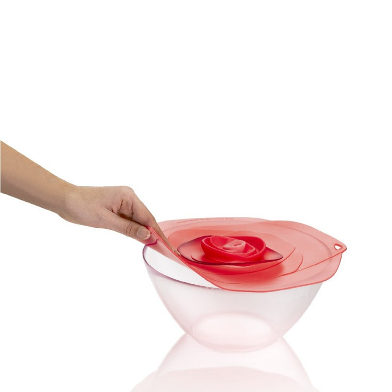 Een hand tilt het Charles Viancin Vershouddeksel Rose Fuchsia aan het lipje van een grote glazen kom