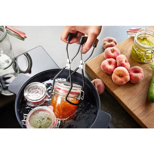 GEFU Glaslifter Lift Up, een hand die met de lifter een gevulde weckpot met oranje jam uit een pan met kokend water tilt, met verse perziken op een houten snijplank ernaast