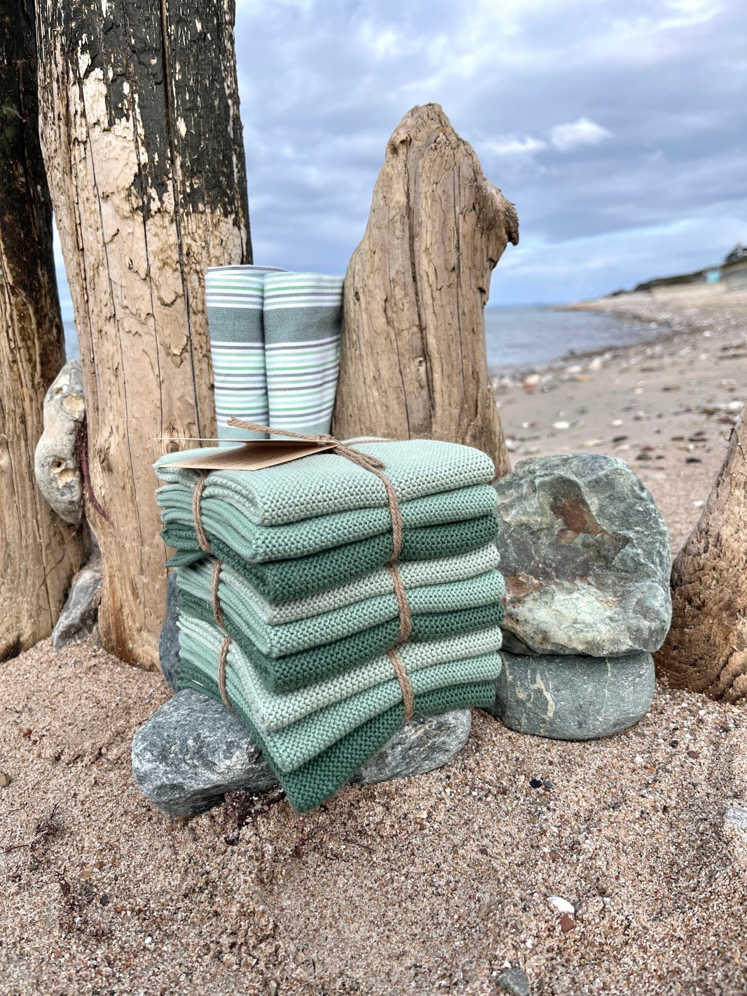 Solwang Design Vaatdoekjes set van 3 - Zeegroen Combi lifestylefoto aan het strand: een gestreept groen-wit-grijs doekje staat opgerold tussen een verweerde houten paal en drijfhout, met daarvoor een stapel zeegroene doekjes samengebonden met touw en een label, tussen stenen op het zand met zee en bewolkte lucht op de achtergrond.