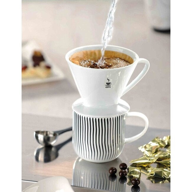 GEFU Koffiefilter Sandro - Maat 1 bovenop een gestreepte mok terwijl er kokend water overheen wordt gegoten, met chocolaatjes op tafel ernaast