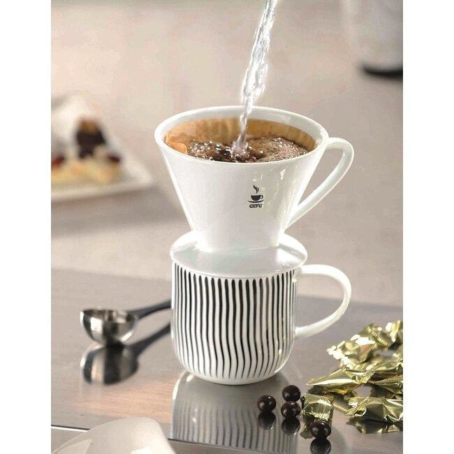 GEFU Koffiefilter Sandro - Maat 2 bovenop een gestreepte mok terwijl er kokend water overheen wordt gegoten, met chocolaatjes op tafel ernaast