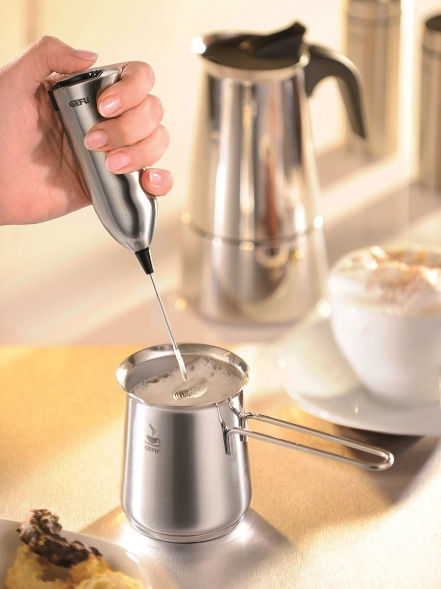 GEFU Opschuimbeker Claudio - 300 ml, hand die met een elektrische melkopschuimer melk in de beker opschuimt naast een percolator