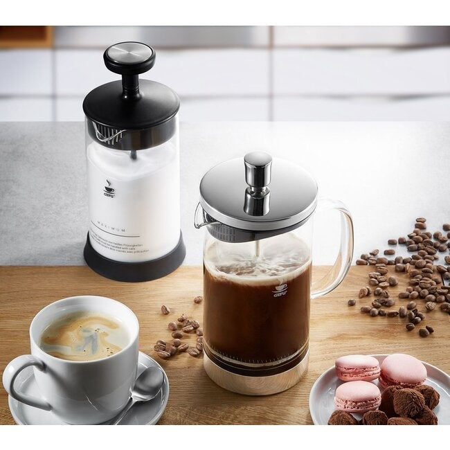 GEFU Cafetière Diego - 1000 ml met zwarte koffie naast een bijpassende melkopschuimer, een kopje koffie, koffiebonen en roze macarons op een houten aanrecht