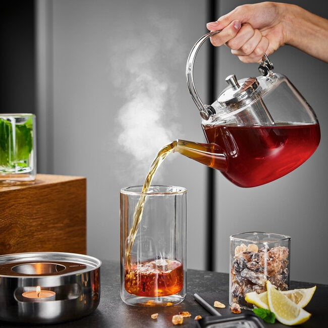 GEFU Theepot Infusa - 1000 ml, een hand schenkt hete thee uit de glazen kan in een glas, met een waxinelichtje eronder, theeblaadjes in een pot en partjes citroen ernaast