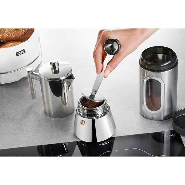 GEFU Koffiestamper met Koffielepel Tamino, close-up van een hand die met de lepel gemalen koffie in een moka-koffiezetter schept op het aanrecht naast een koffieblik en een broodmand