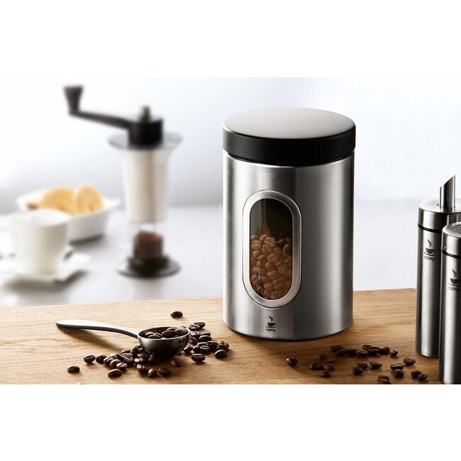 GEFU Koffieblik Piero - 500 g op een houten aanrecht naast een handmatige koffiemolen, een kopje koffie met koekjes en een lepel met gemalen koffiebonen
