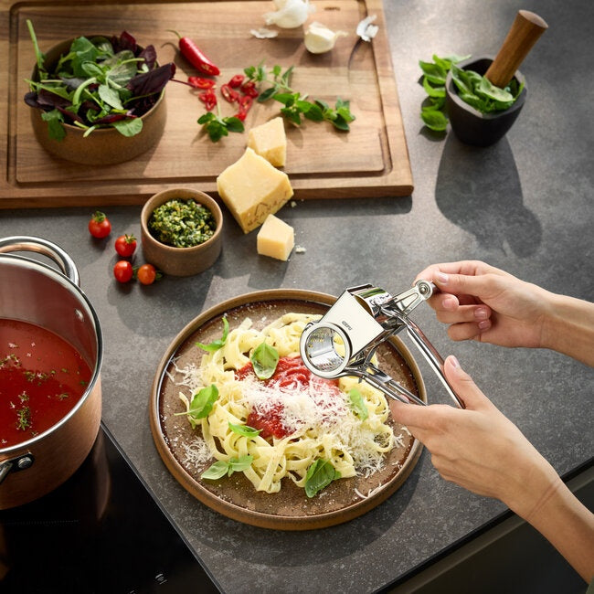 GEFU Kaasmolen Queso - RVS, een hand die met de molen kaas raspt boven een bord tagliatelle, met een snijplank met kruiden en kaas op de achtergrond