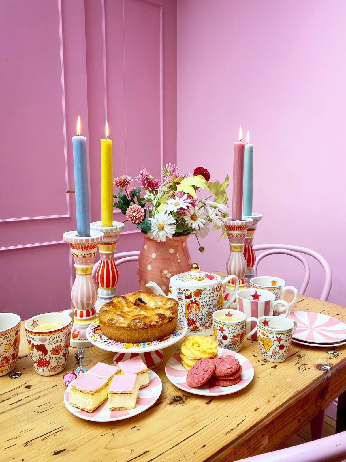 Blond Amsterdam servies op gedekte tafel met taart, koekjes en gekleurde kaarsen tegen roze muur.