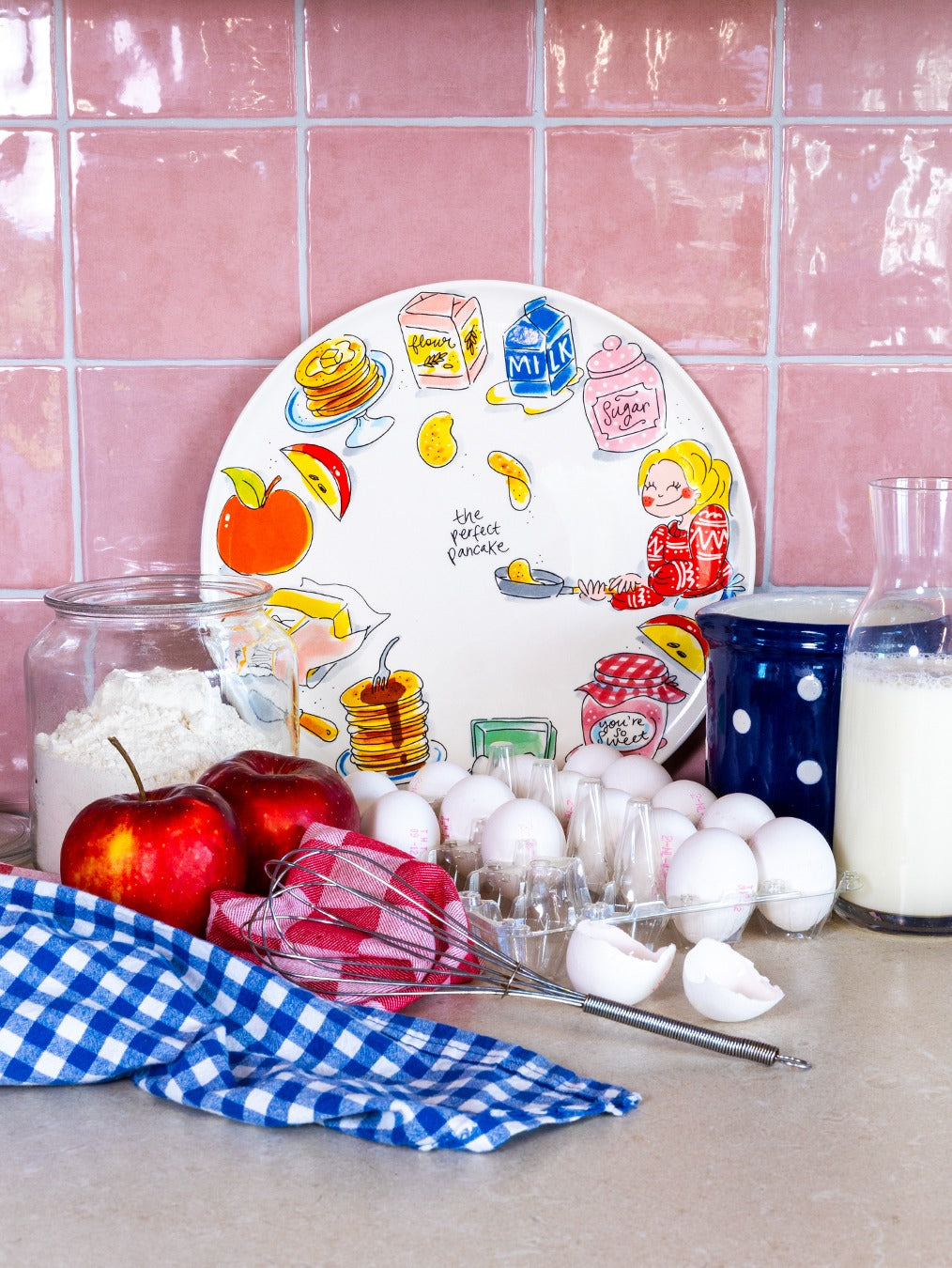 Blond Amsterdam Perfect Pancake serveerbord tegen een roze tegelwand, met appels, bloem, eieren en een garde ernaast