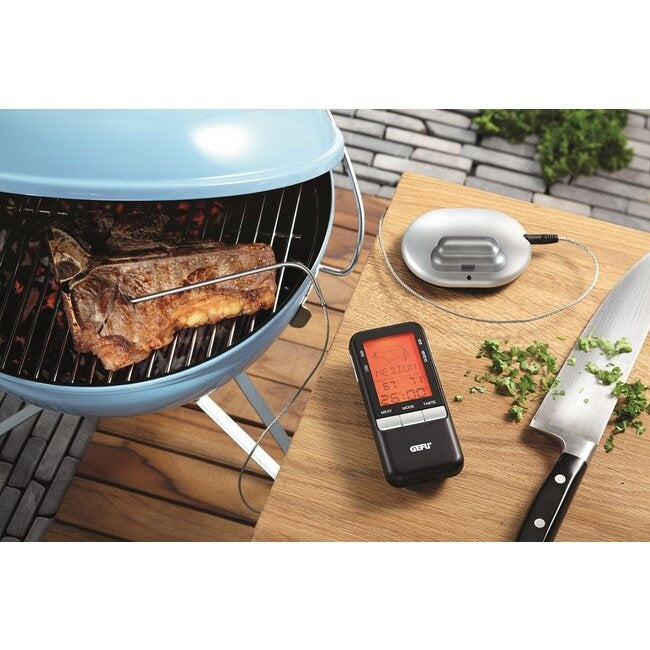 GEFU Digitale Thermometer Handi op een blauwe kogelgrill met een geheel stuk vlees op de grill, de zwarte display-eenheid met kabel op een houten snijplank naast een mes en verse kruiden