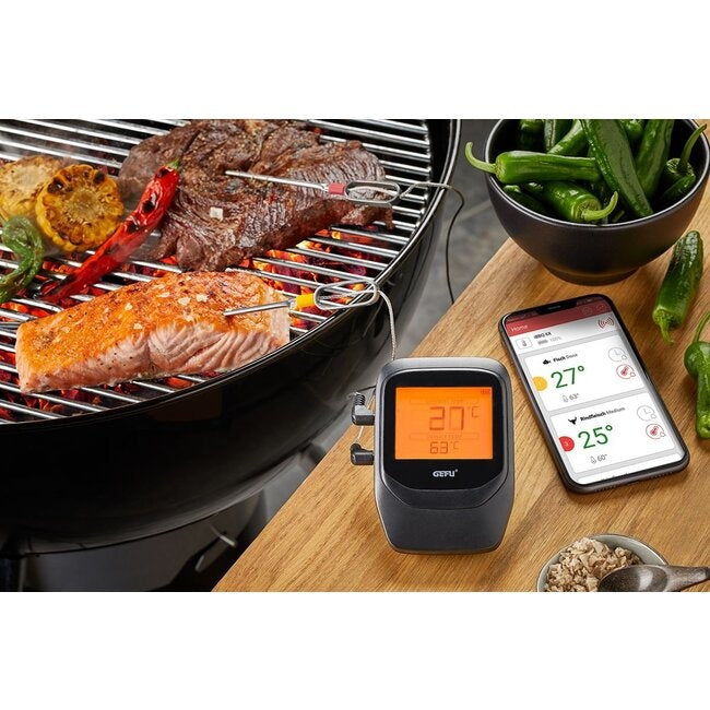 GEFU Grill- en Braadthermometer Control - 6 Kanalen, twee meetpennen in zalm en biefstuk op de grill, met de display-eenheid en een smartphone met de app die twee temperaturen toont op een houten tafel