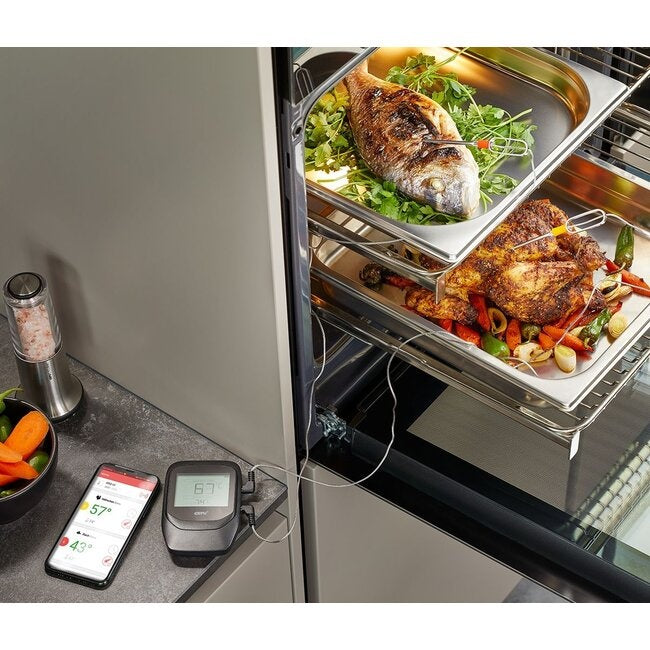 GEFU Grill- en Braadthermometer Control - 6 Kanalen bij een oven met bakplaten vol geroosterde vis en kip, de display-eenheid en smartphone met app op het aanrecht ernaast
