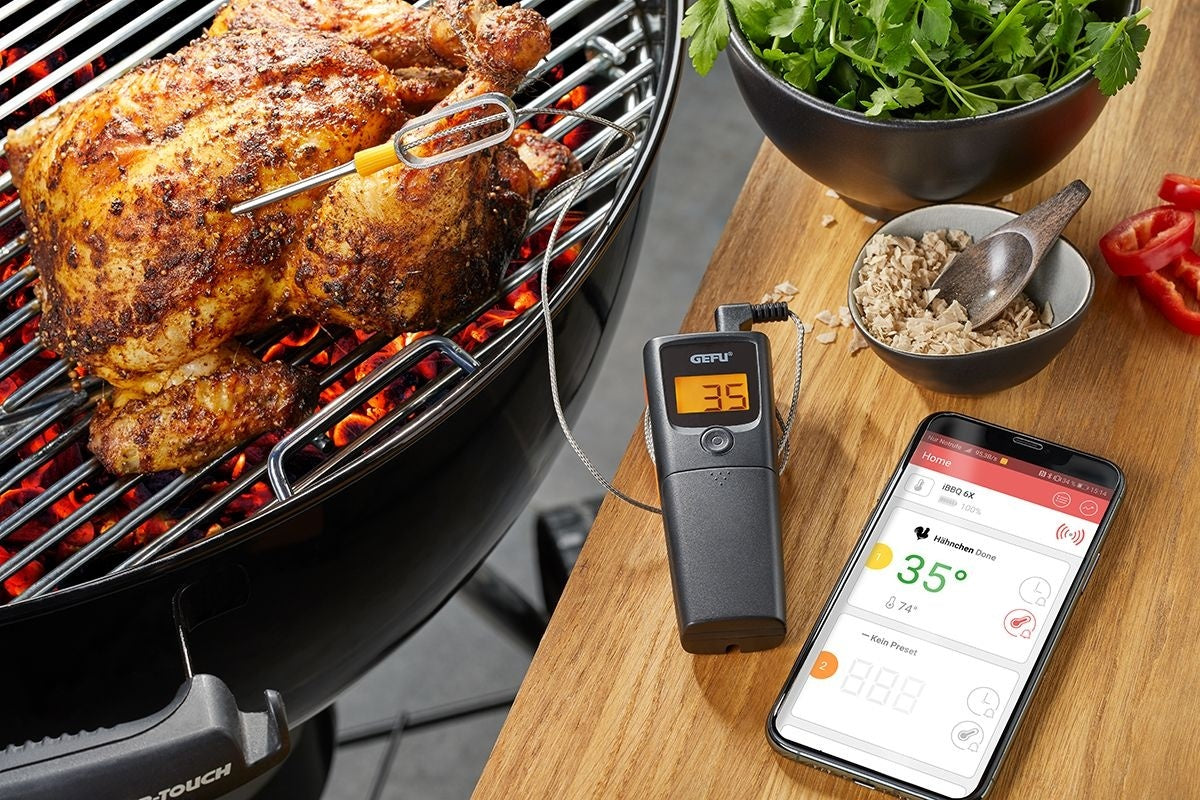 GEFU Grill- en Braadthermometer Control - 1 Kanaal, de meetpen in een geheel gebraden kip op de barbecue, met de display-eenheid en een smartphone met de bijbehorende app op een houten tafel ernaast