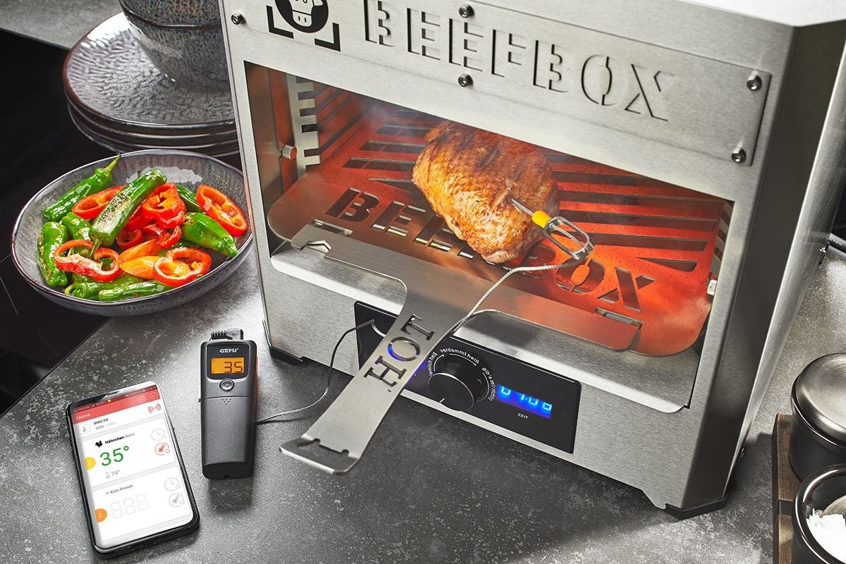 GEFU Grill- en Braadthermometer Control - 1 Kanaal naast een rotisserie-oven met vlees aan het spit, de display-eenheid en smartphone met app op het aanrecht bij schaaltjes met groente
