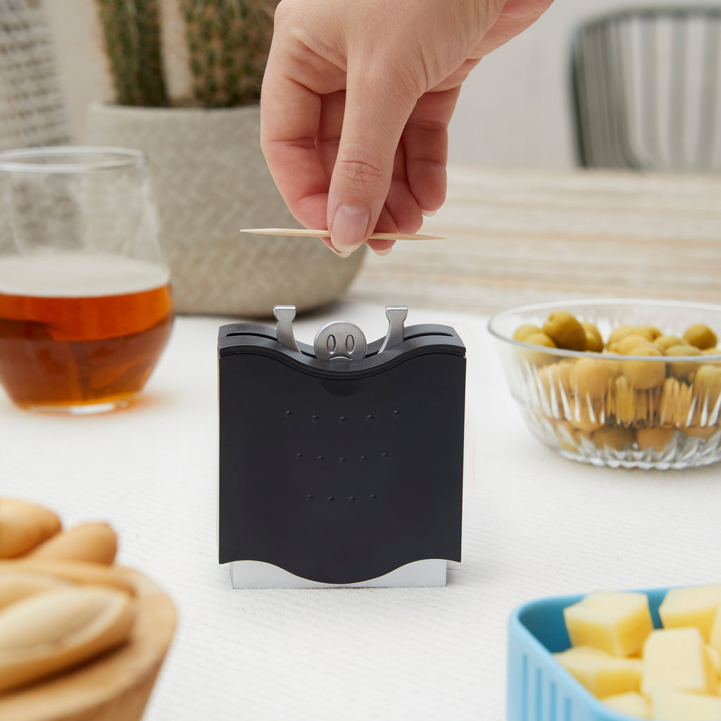 Balvi tandenstokerdispenser op tafel met bier, olijven en kaasblokjes, hand pakt een tandenstoker
