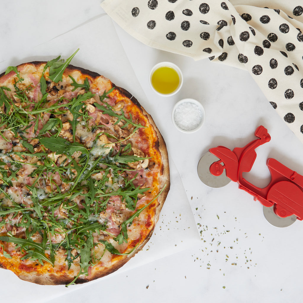 Pizza met rucola en ham naast de rode Balvi scooter-pizzasnijder, met olie en zout