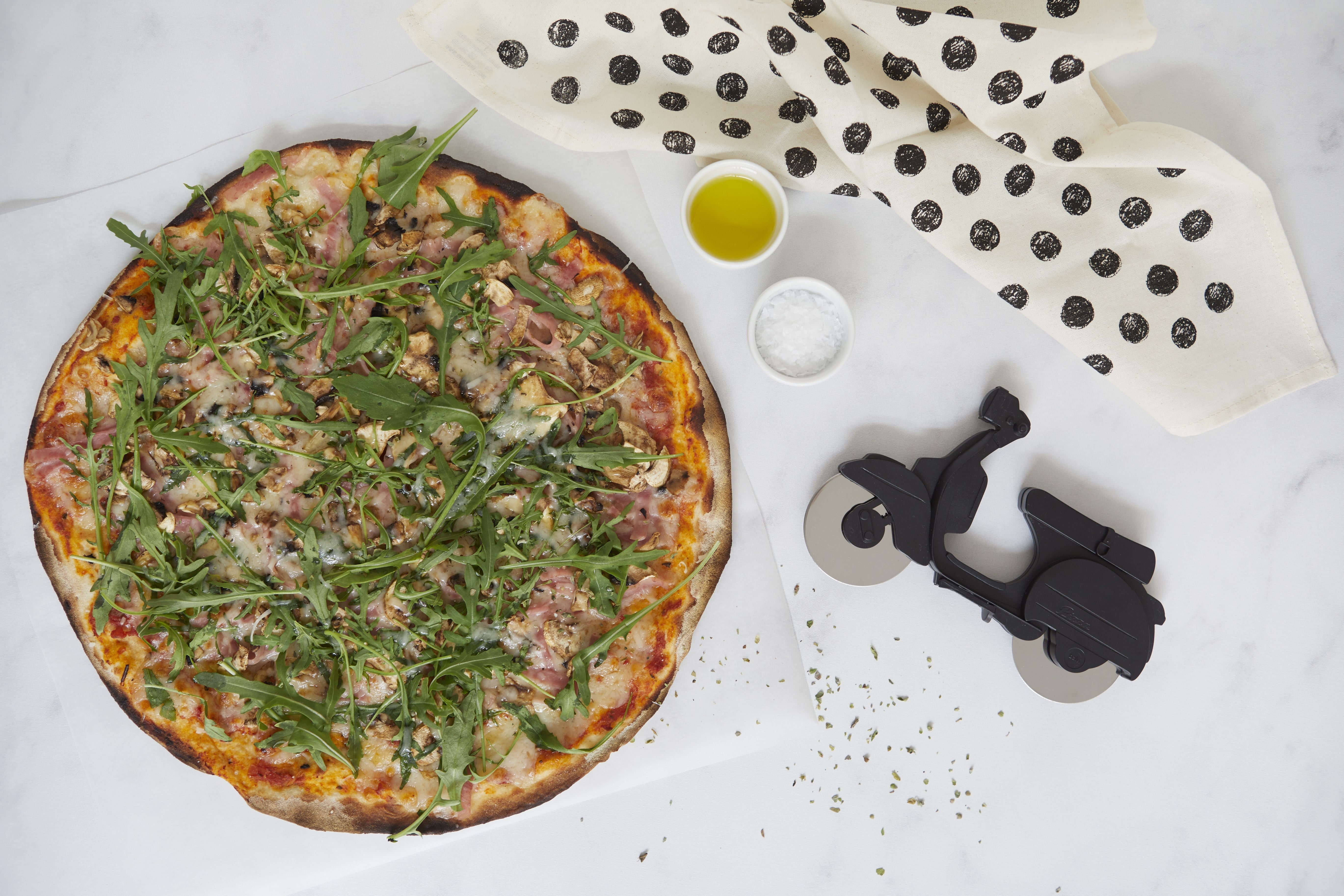 Balvi Pizzasnijder Scooter Zwart, op een gestippeld werkblad naast een pizza