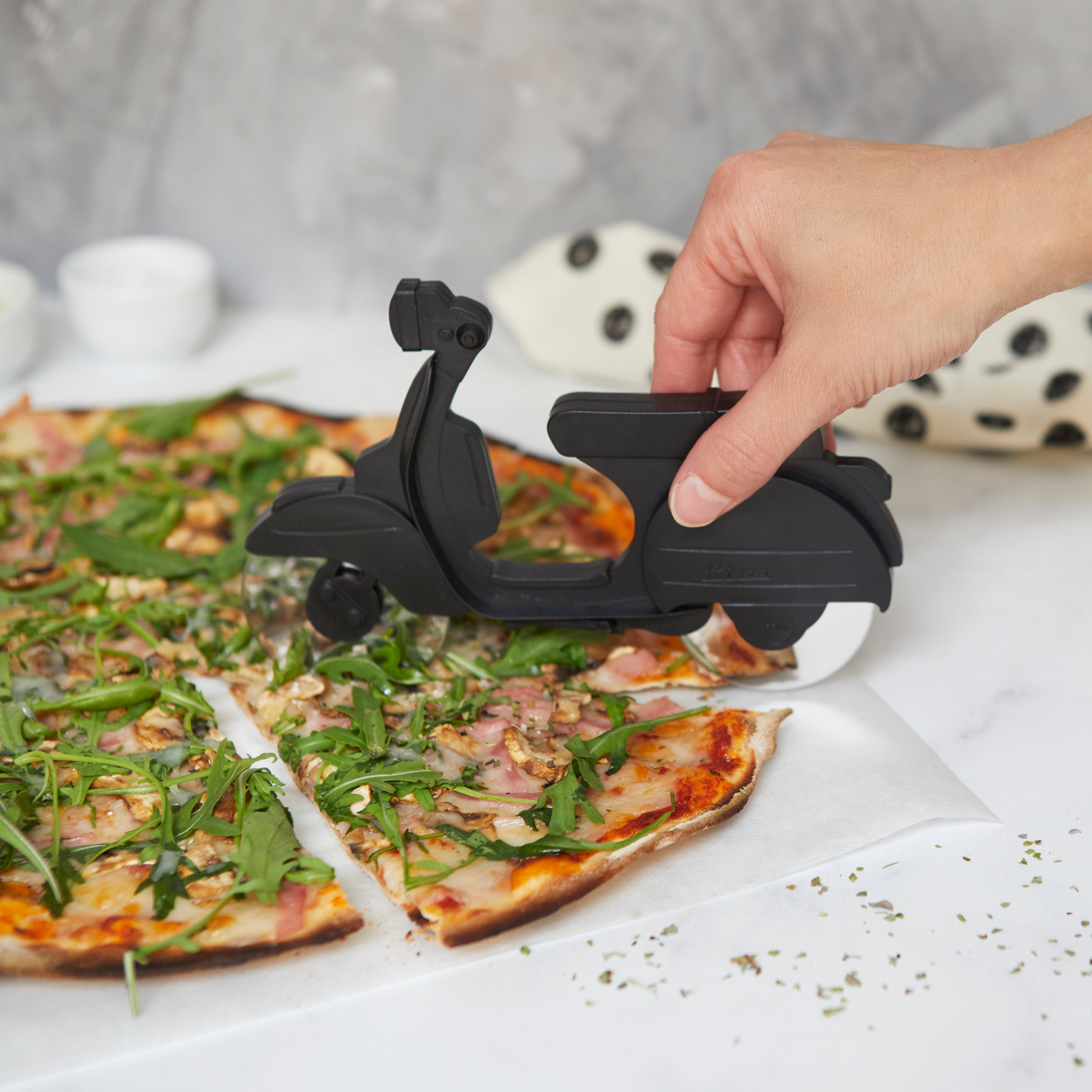 Balvi Pizzasnijder Scooter Zwart, hand rijdt de scooter over een pizza om 'm te snijden