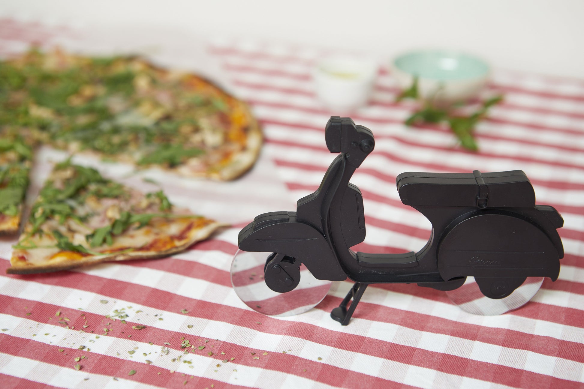 Balvi Pizzasnijder Scooter Zwart, op een rood-wit geblokt tafelkleed naast een pizza