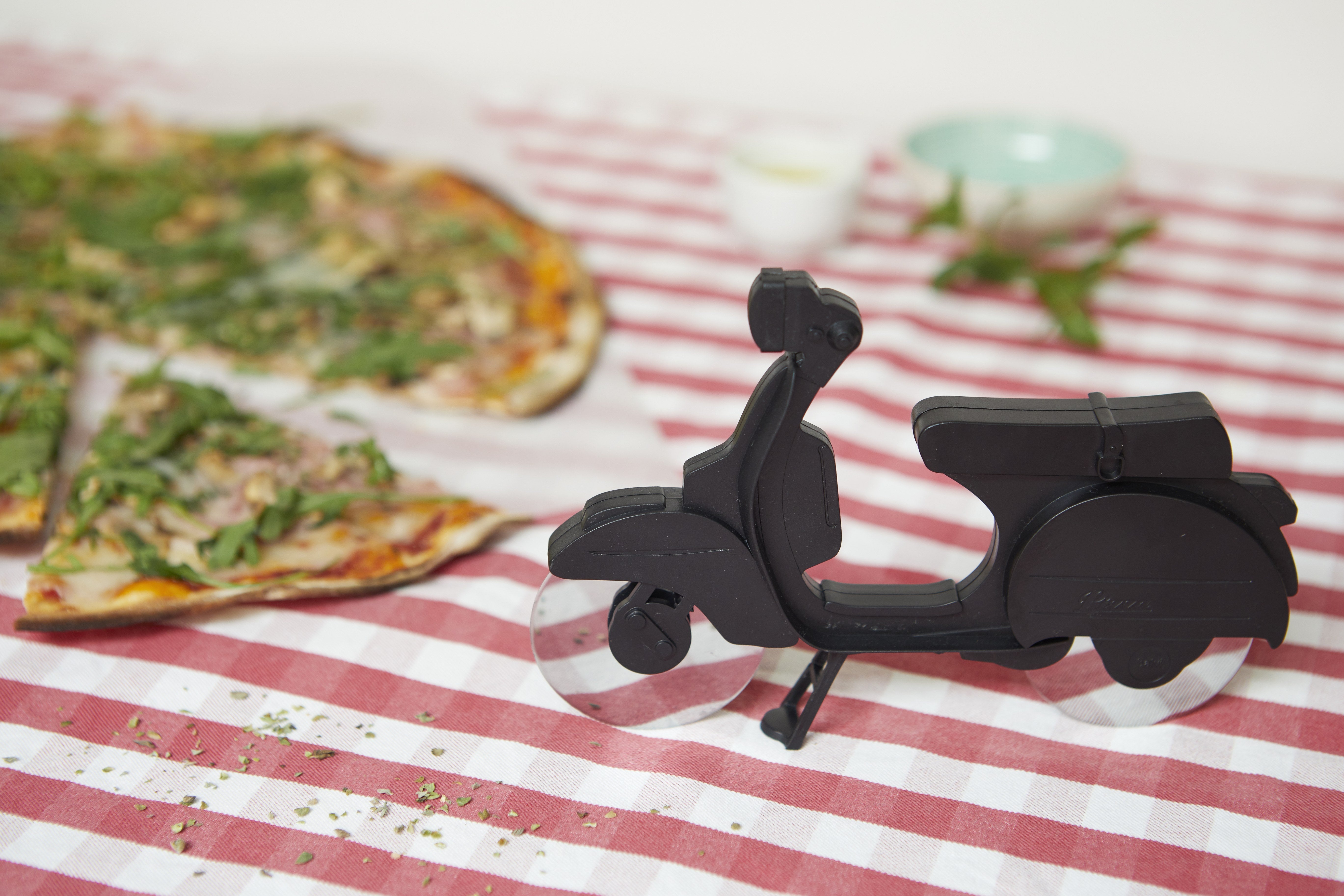 Balvi Pizzasnijder Scooter Zwart, op een rood-wit geblokt tafelkleed naast een pizza