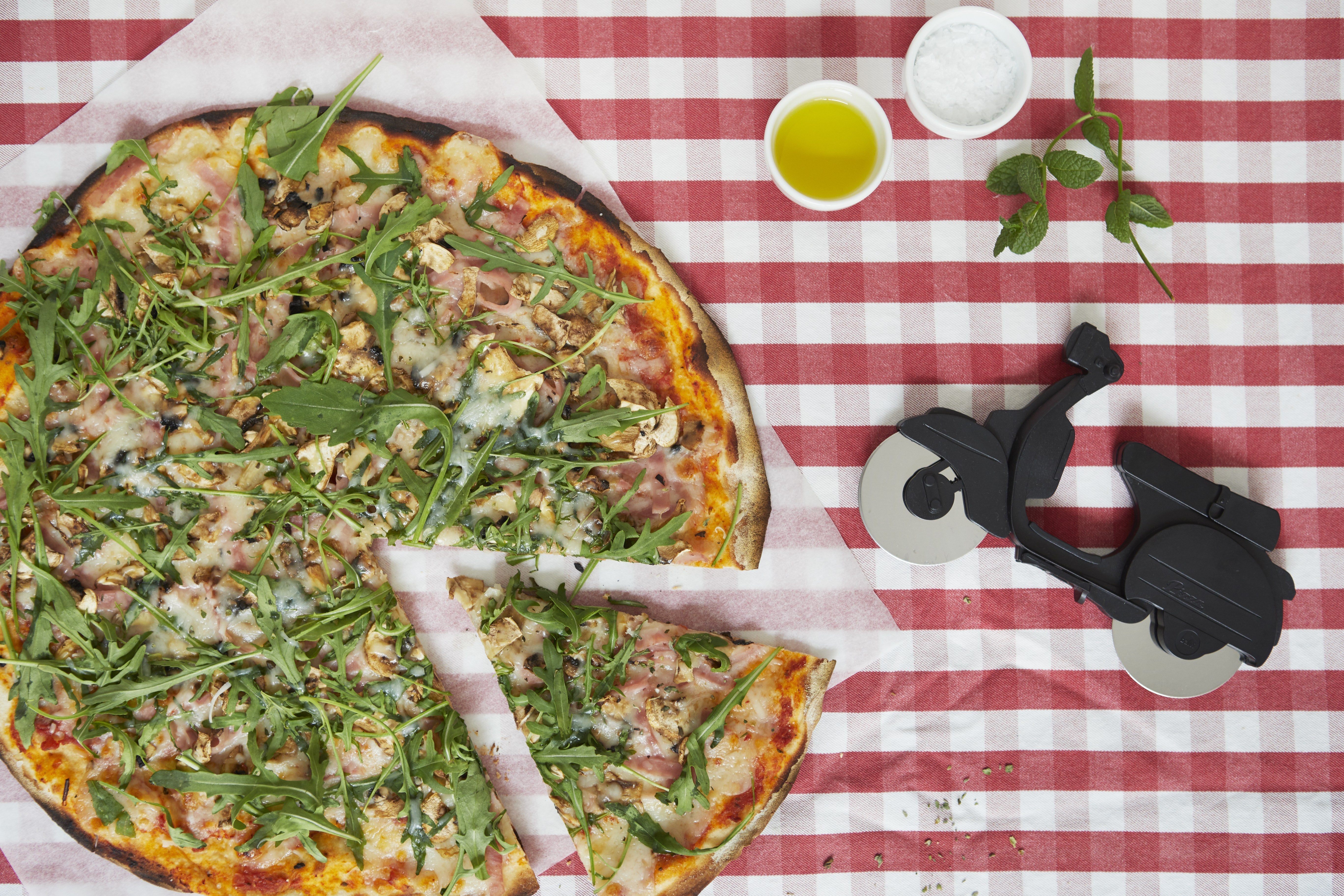Balvi Pizzasnijder Scooter Zwart, op een rood-wit geblokt tafelkleed, close-up van een aangesneden pizzapunt