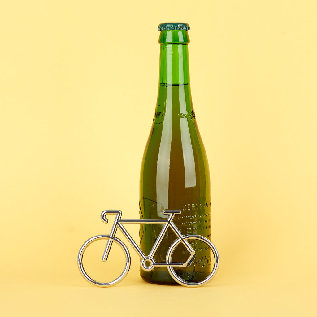 Zilverkleurige fietsvormige flesopener liggend naast een groene bierfles tegen een gele achtergrond