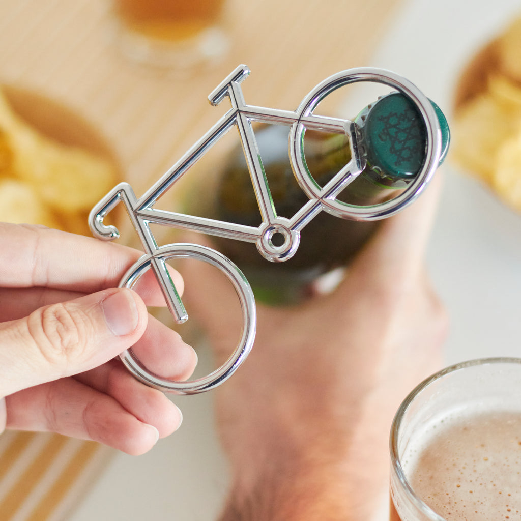Hand houdt de zilverkleurige fietsopener op de kroonkurk van een flesje, met chips en een glas bier onscherp op de achtergrond