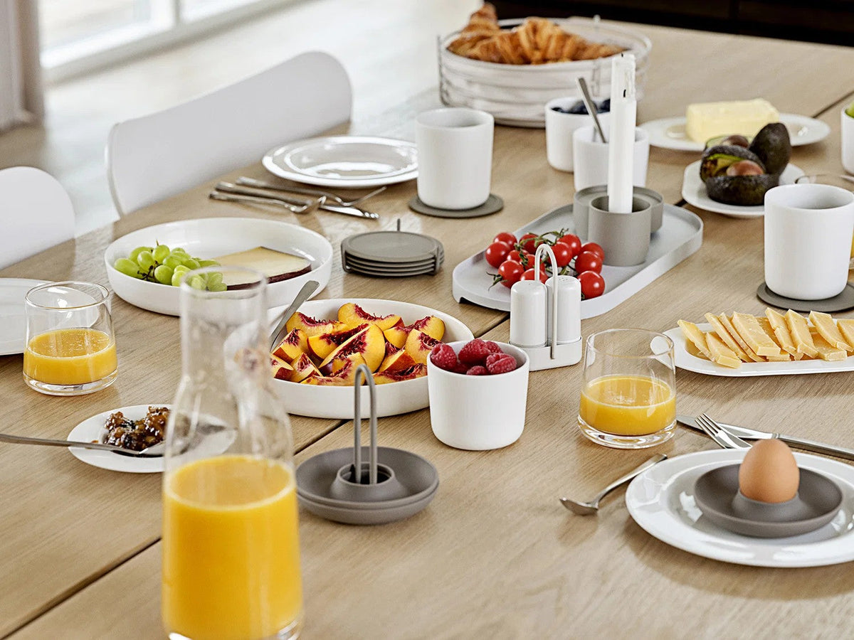 Gedekte eikenhouten tafel met wit servies, glazen sinaasappelsap en een stapel taupe onderzetters in het midden