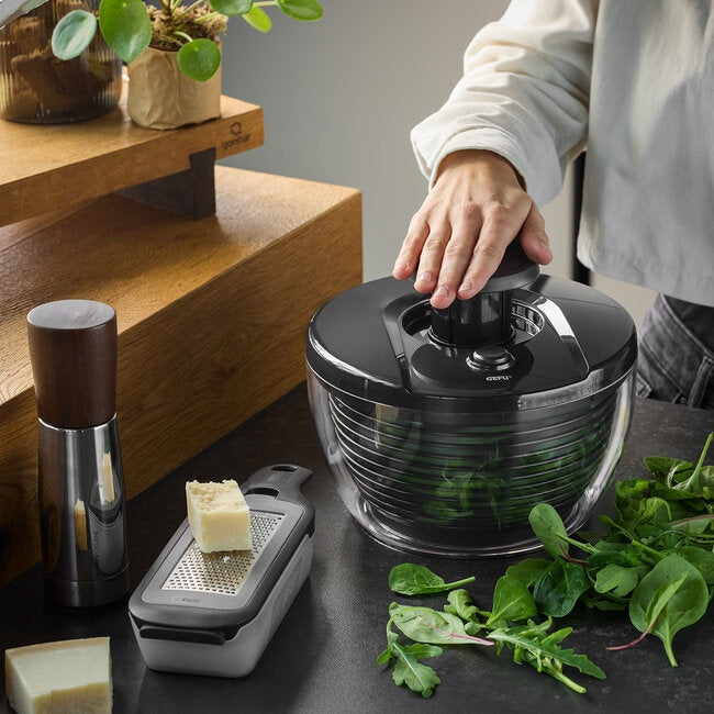 GEFU Saladespinner Push It - 3 liter, een hand drukt op de zwarte pushknop op het aanrecht naast een rasp met geraspte kaas en verse rucola