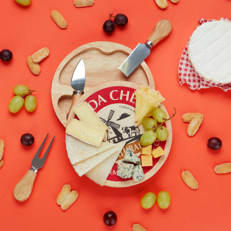 Plank met opdruk Gouda Cheese gevuld met kaasplakken, blauwe kaas en druiven op een oranje achtergrond naast de mesjes