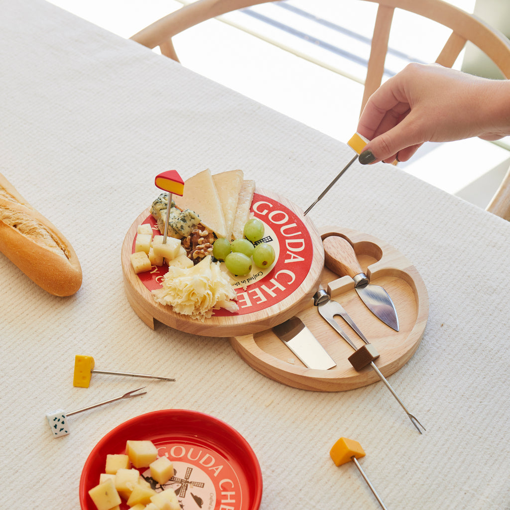 Kaasplank op een gedekte tafel met opengeschoven mesjesvak, kaasjes op prikkers en een rood schaaltje