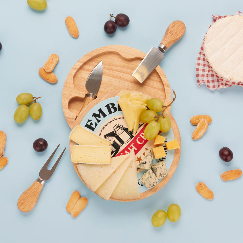 Plank met Camembert-opdruk met kaasplakken, blauwe kaas en druiven op een lichtblauwe achtergrond naast de mesjes