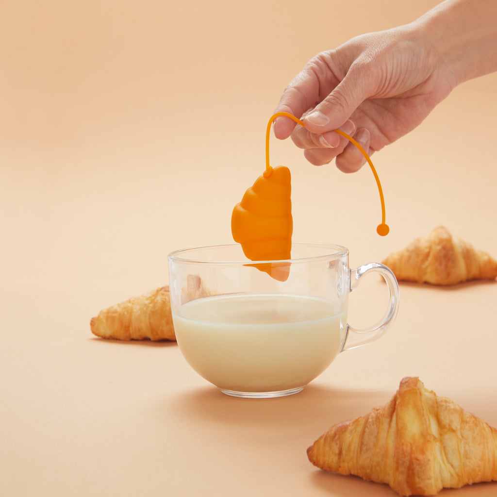 Hand houdt het croissant-thee-ei aan het koord boven een glazen kop, met croissants op een perzikkleurige achtergrond
