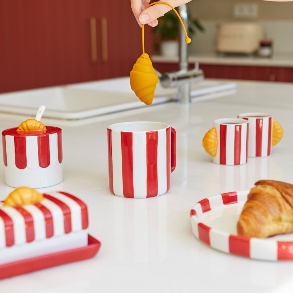 Croissant-thee-ei aan het koord boven een wit aanrecht met rood-wit gestreepte kopjes en een bord met een croissant