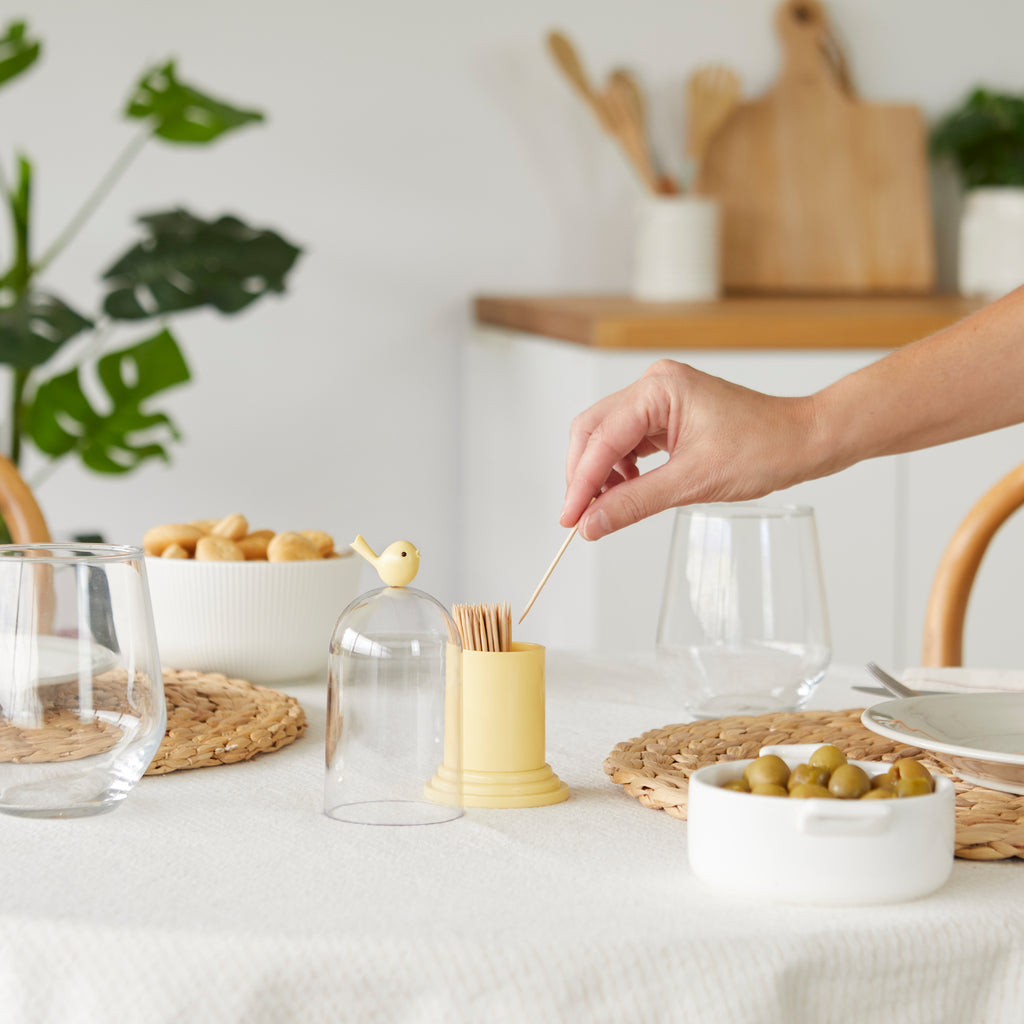 Hand pakt een tandenstoker uit de gele Birdie op een gedekte tafel naast olijven en een kom noten