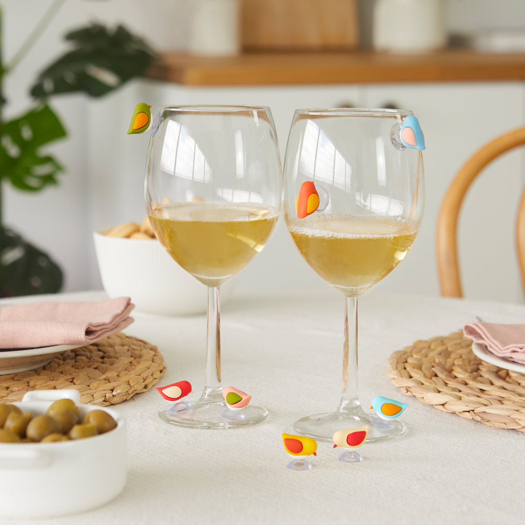 Gedekte tafel met twee glazen witte wijn, vogelmarkers op de glazen en op het tafelkleed, naast een schaaltje olijven