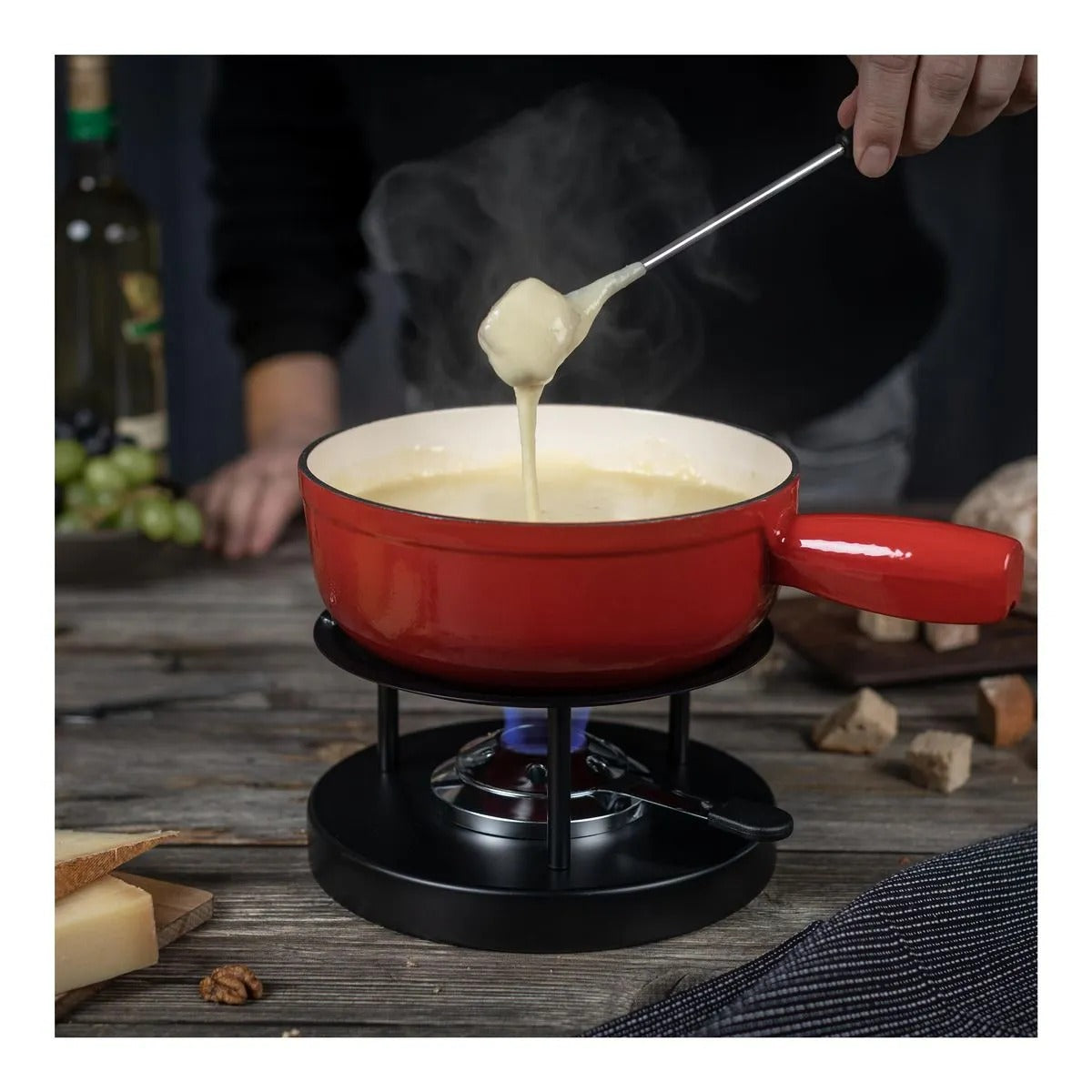 Hand doopt een fonduevork met kaas boven de rode 24 cm Caquelon, met wijn, druiven en kaas op de achtergrond.